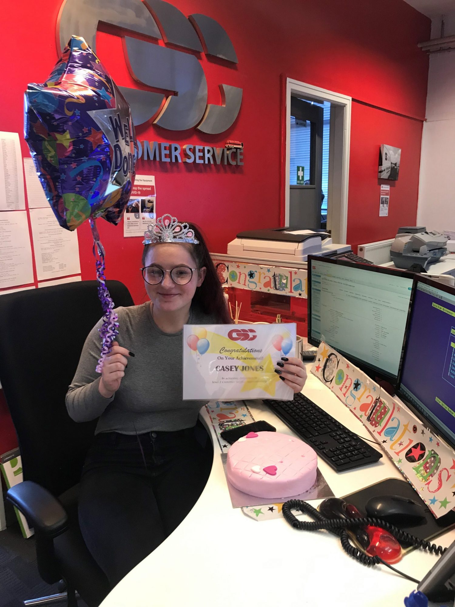 Casey Jones celebrating achieving Distinction on her Apprenticeship, with cake, balloons and a diploma